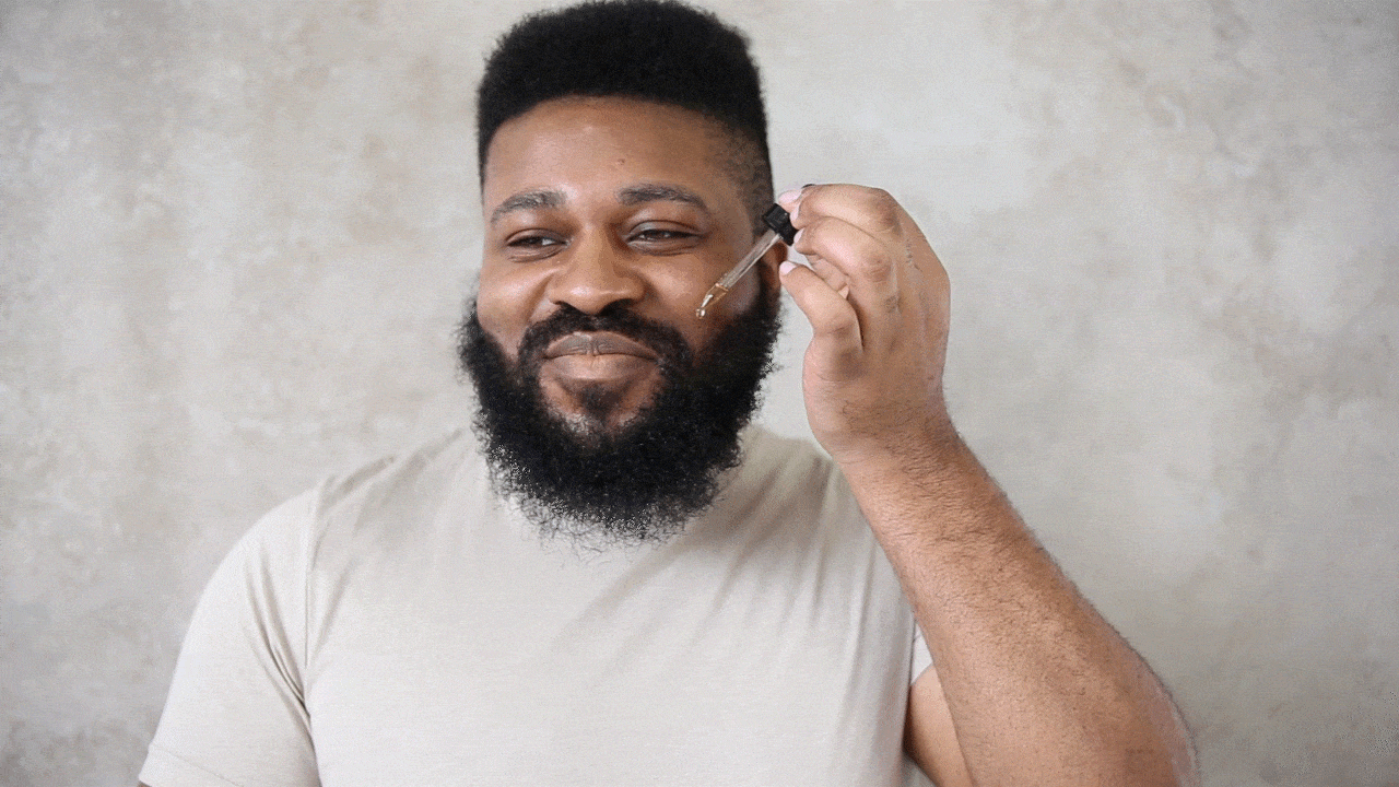 Man smiles as he applies beard oil with a dropper.