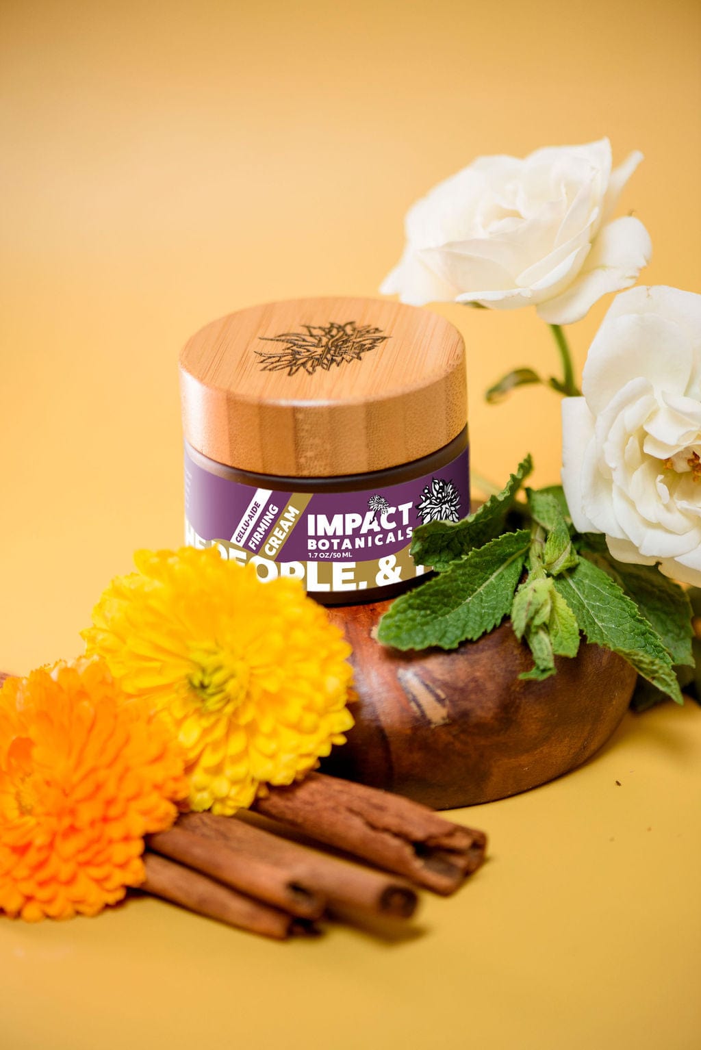 A jar of Impact Botanicals Cellulite Firming Cream sits on a wooden surface surrounded by vibrant marigolds, white roses, fresh mint, and cinnamon sticks.