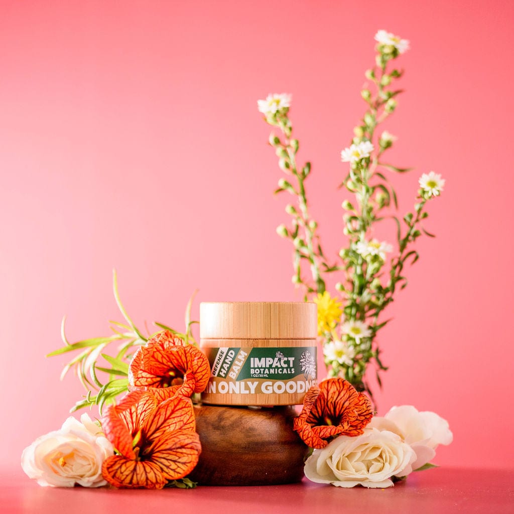 Bamboo jar of Defense Hand Balm by Impact Botanicals surrounded by vibrant orange and white flowers on a pink background.