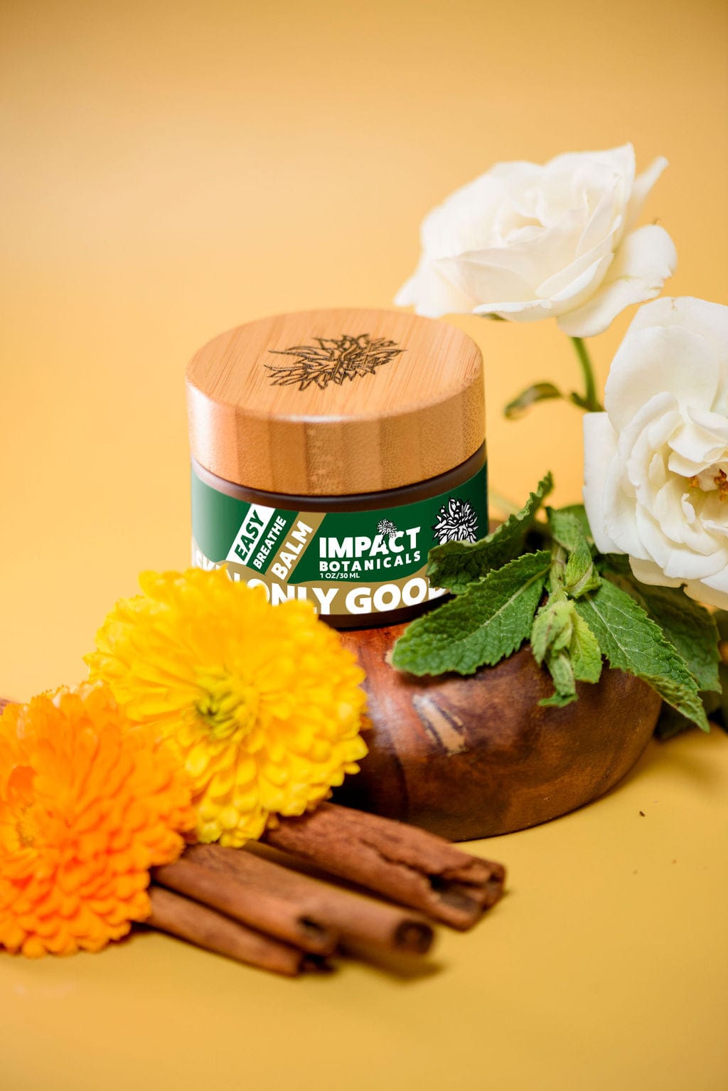 An amber jar of Impact Botanicals Easy Breathe Balm rests on a wooden surface. White roses, vibrant calendula flowers, and sprigs of mint surround the balm, hinting at its calming and soothing aromatherapy properties.