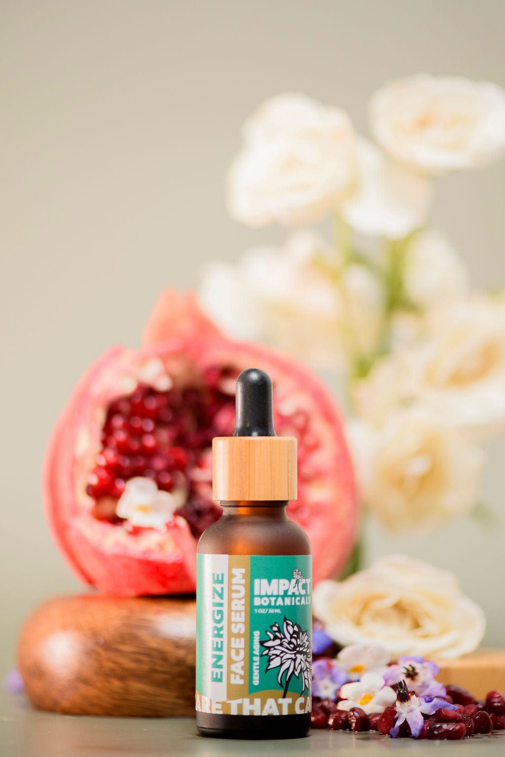 A bottle of Energize Face Serum sits on a wooden platform with a halved pomegranate behind it and assorted flowers scattered around.