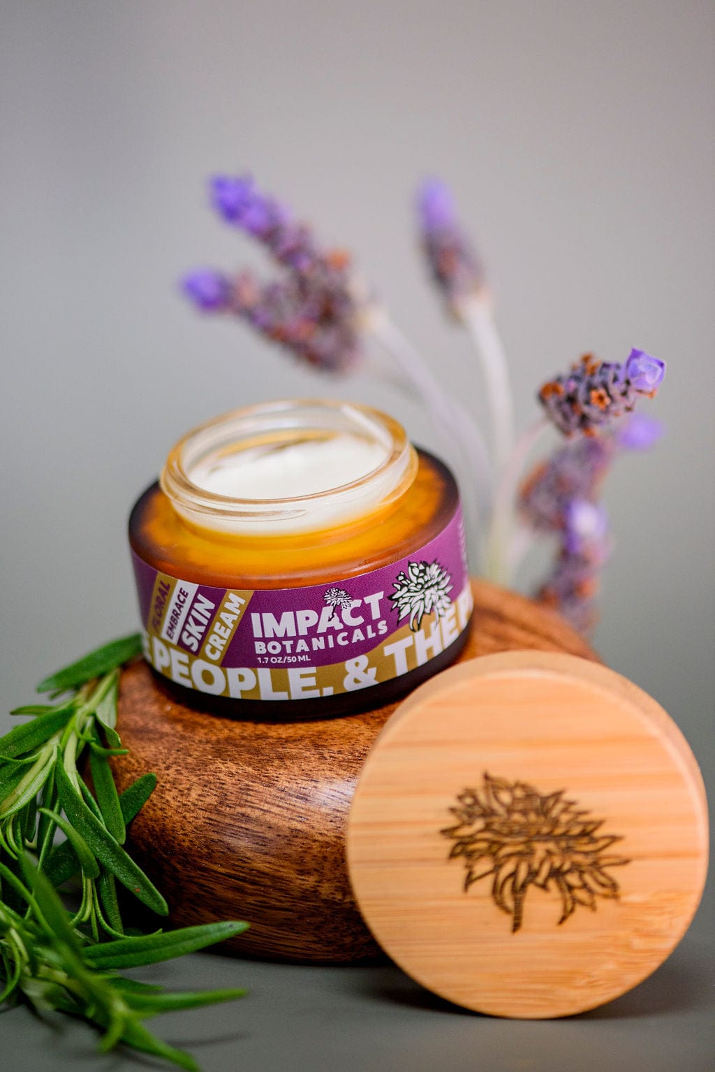 Open jar of Impact Botanicals face cream sits on a wooden platform with the bamboo lid beside it. Sprigs of lavender and rosemary surround the moisturizer.
