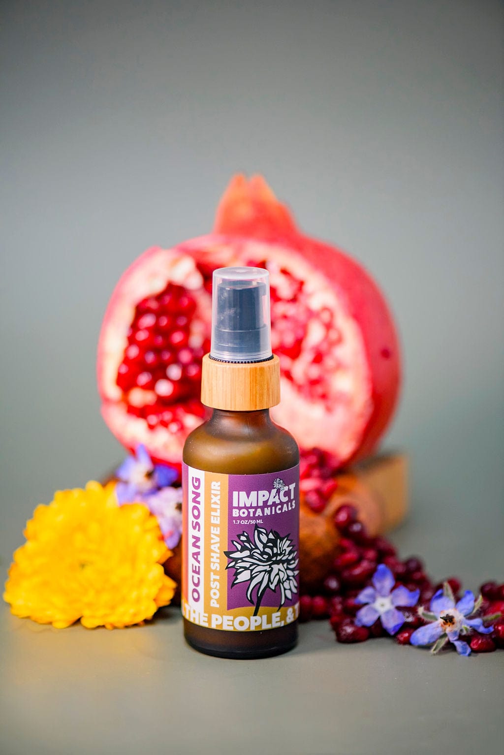 Impact Botanicals Ocean Song Post Shave Elixir bottle stands beside a halved pomegranate and vibrant flowers.