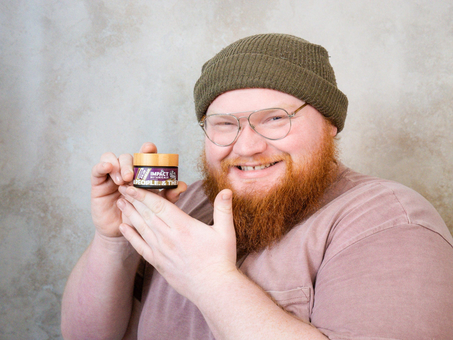 Redheaded man with a beard and glasses smiles while holding a jar of Impact Botanicals skin cream.