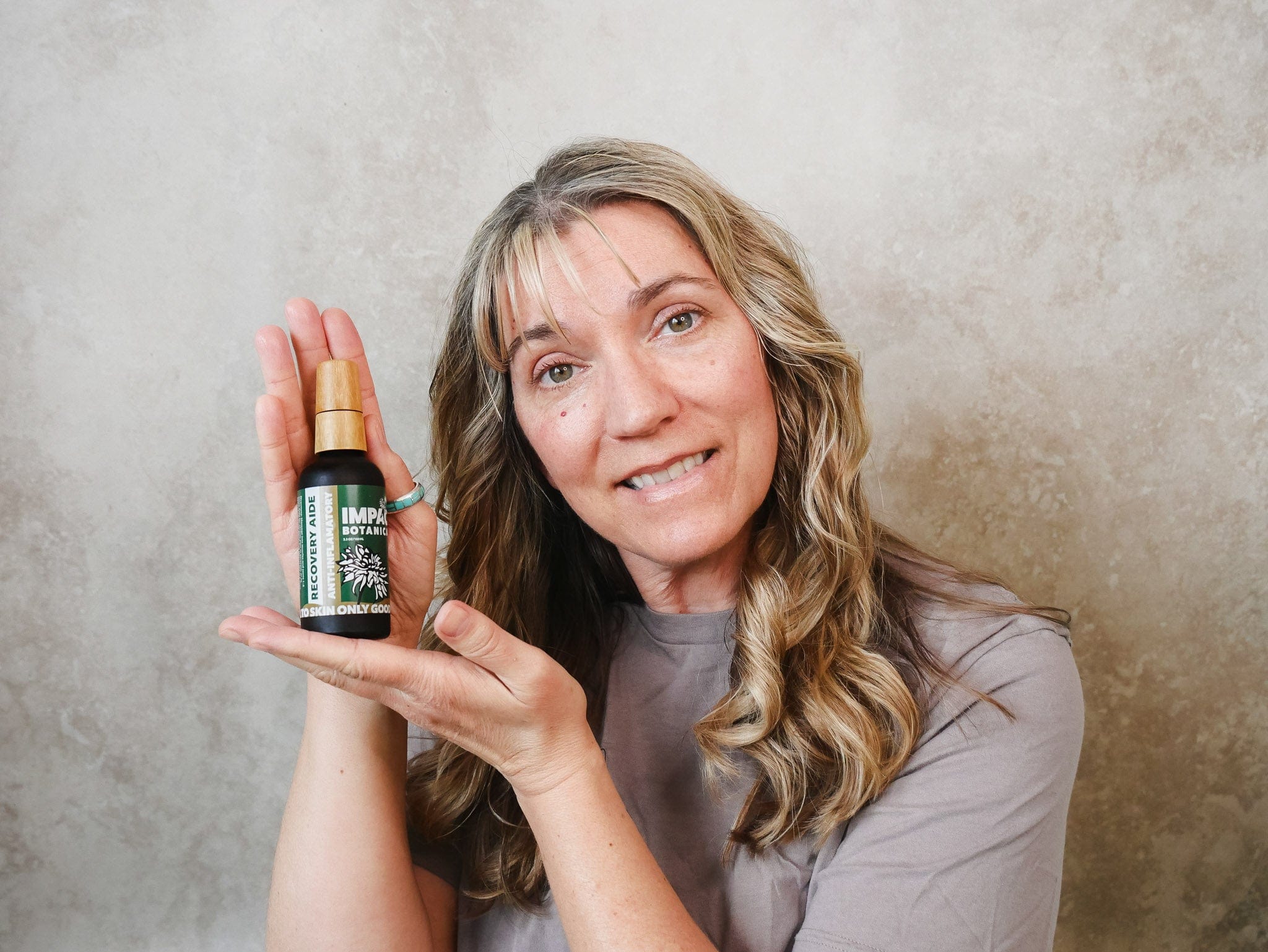 Woman smiles while holding a bottle of Impact Botanic Recovery Aide anti-inflammatory serum.