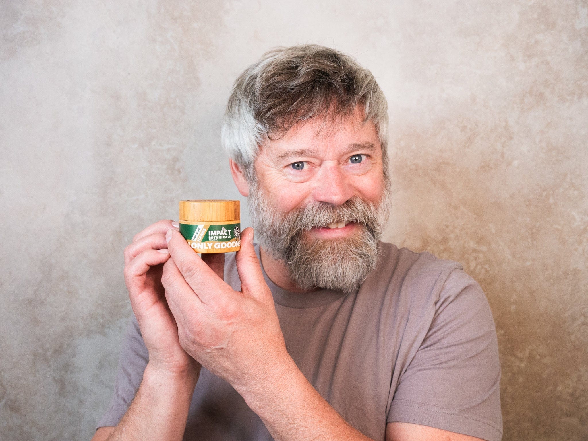 Smiling man holds a jar of Impact Botanicals Working Hands cream.