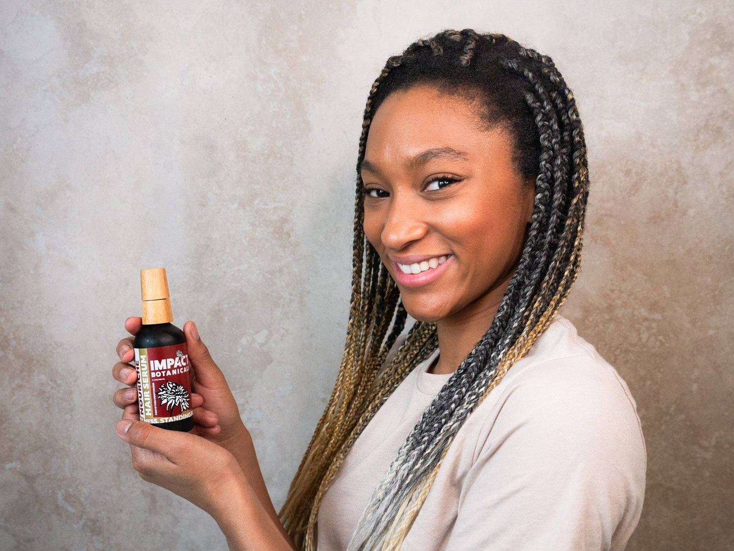 Woman smiles as she holds a bottle of Nourish Hair Serum by Impact Botanicals.