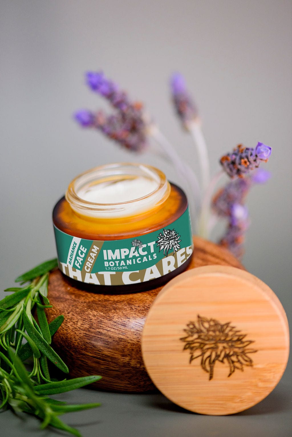 Open jar of Impact Botanicals Radiance Face Cream sits on a wooden display beside its bamboo lid and sprigs of lavender and rosemary.