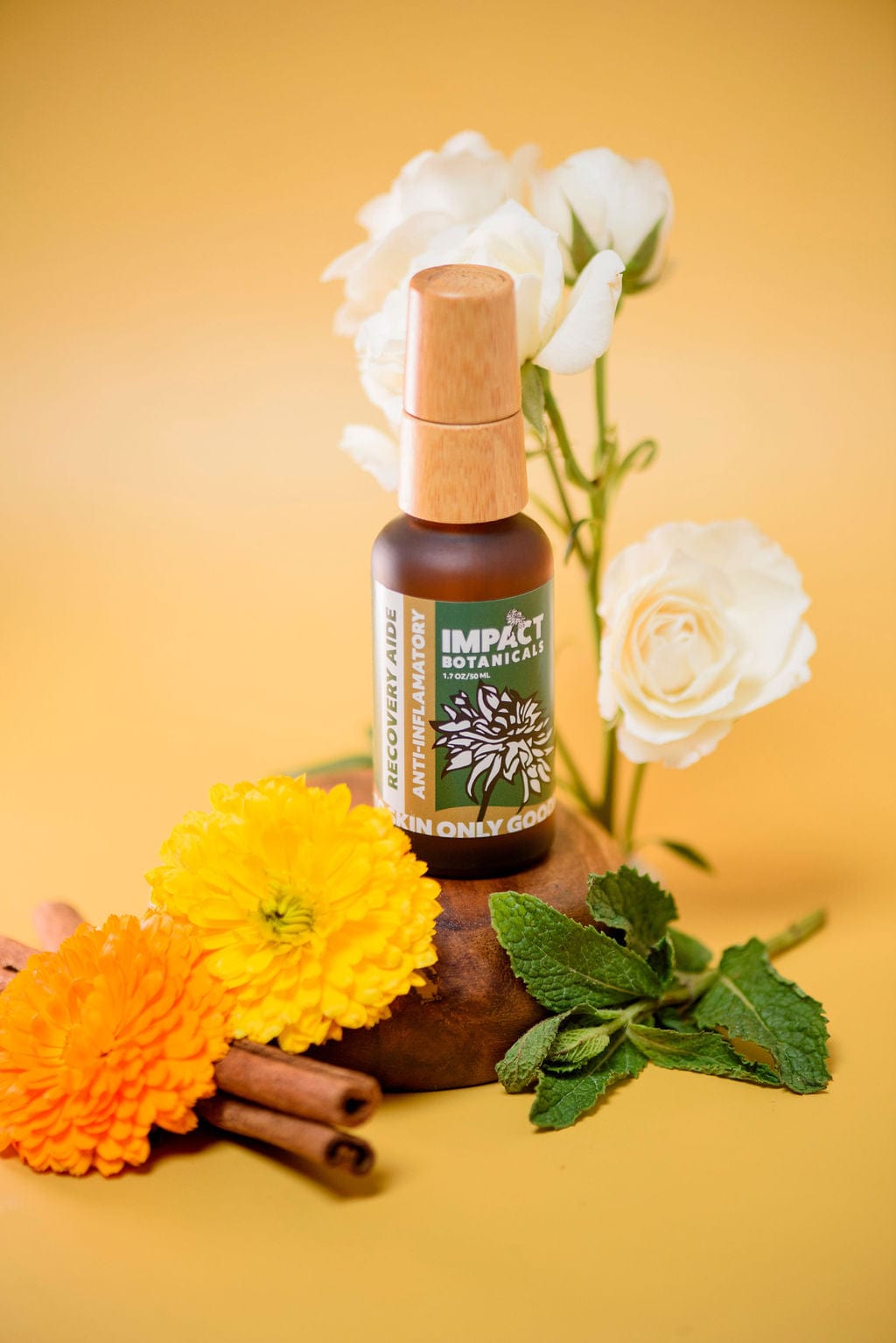 A bottle of Impact Botanicals Recovery Aide anti-inflammatory serum stands on a wooden platform surrounded by white roses, bright orange calendula flowers, cinnamon sticks, and fresh mint leaves.