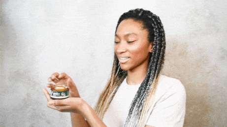 Woman with long braids smiles as she holds a small jar of skincare product.