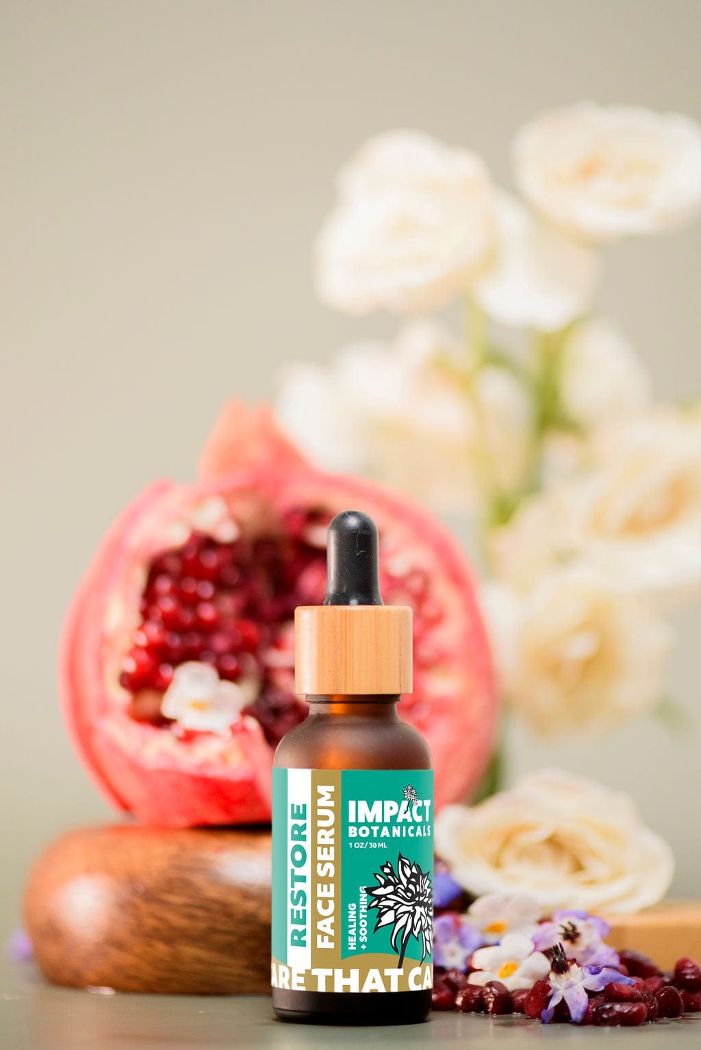 A bottle of Impact Botanicals Restore Face Serum sits on a wooden surface with a halved pomegranate behind it and white flowers in the background.