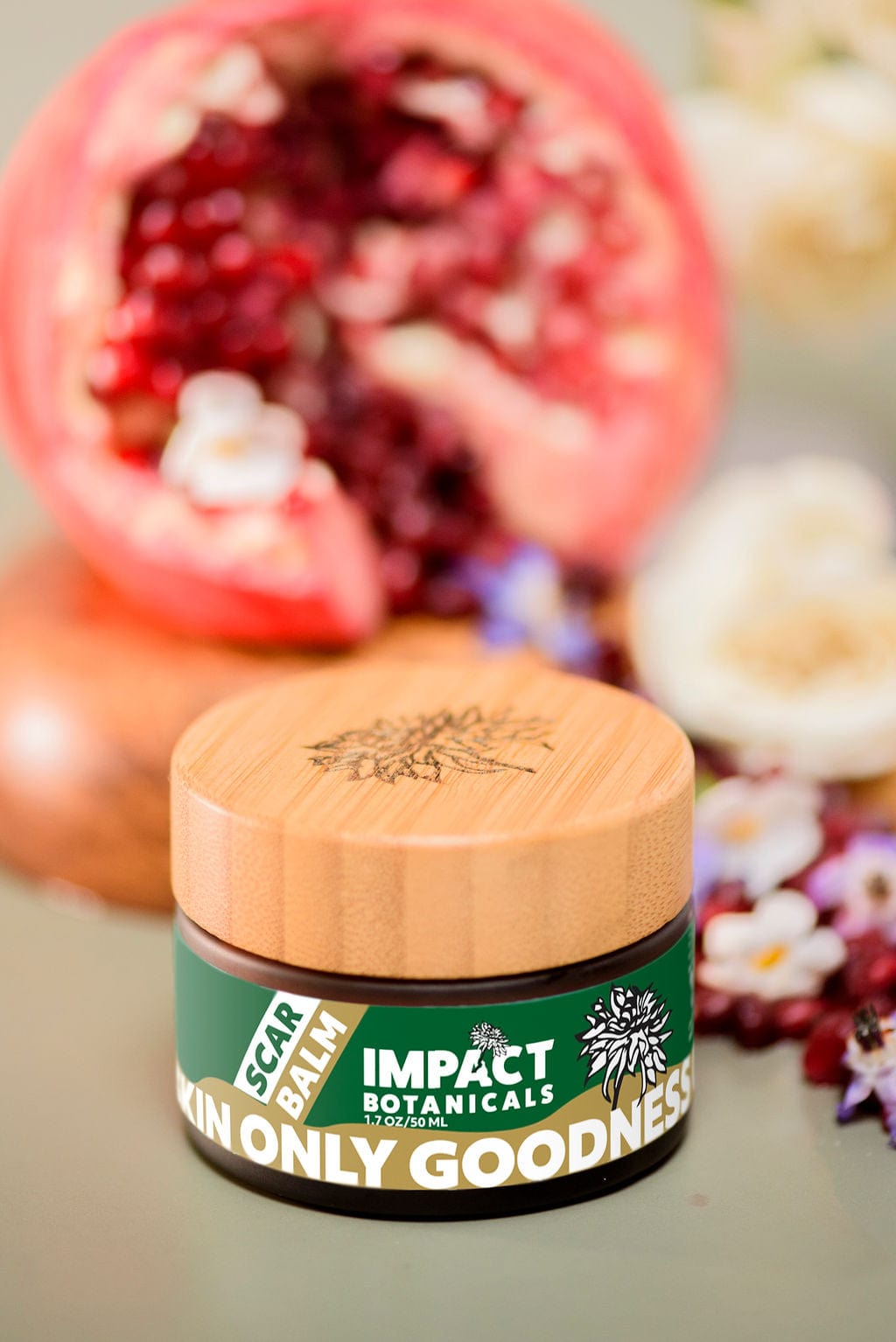 Impact Botanicals Scar Balm in glass jar with bamboo lid, surrounded by fresh pomegranate and delicate flowers.