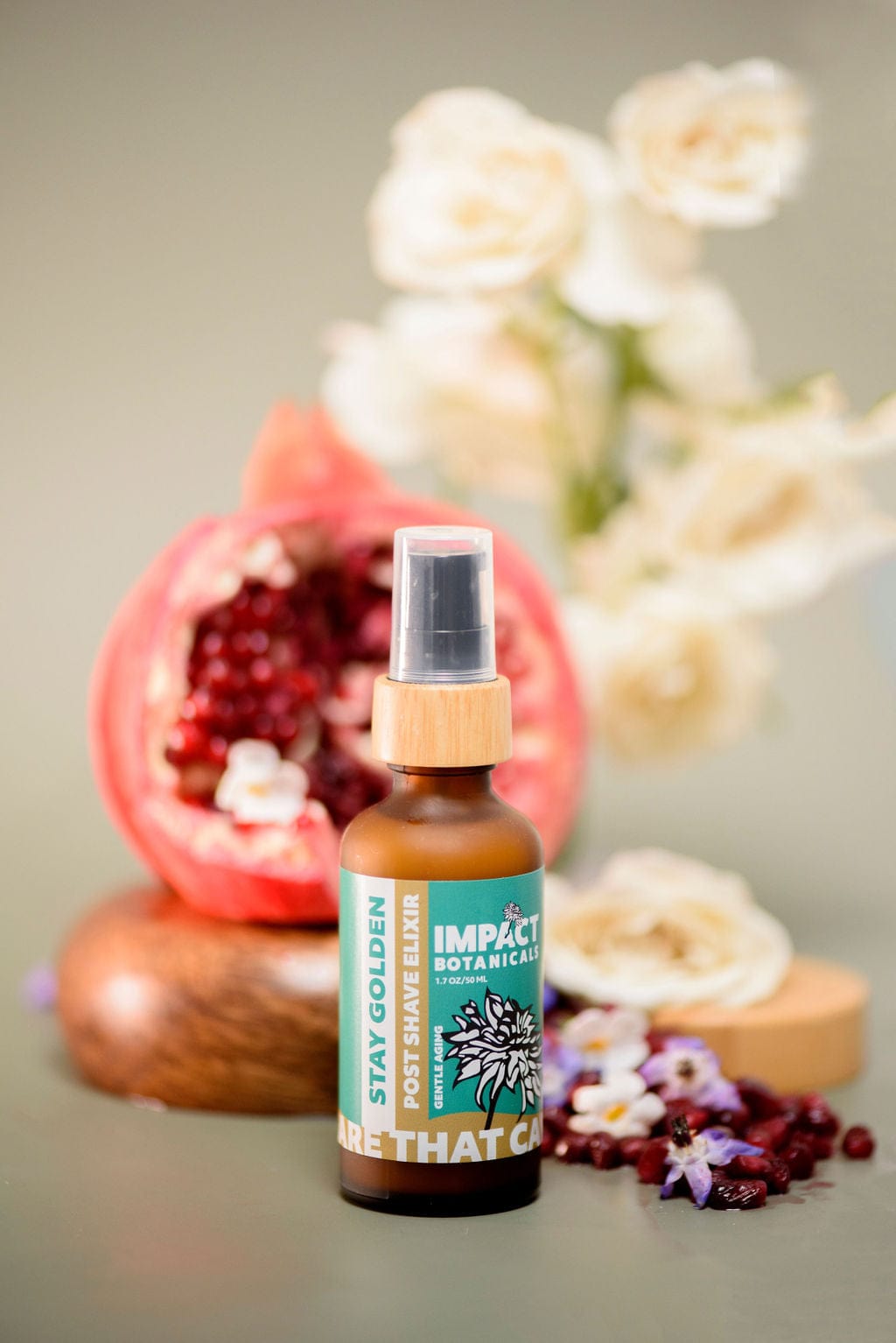 "Stay Golden Post Shave Elixir" bottle by Impact Botanicals with a bamboo lid stands beside a halved pomegranate and scattered flower petals.