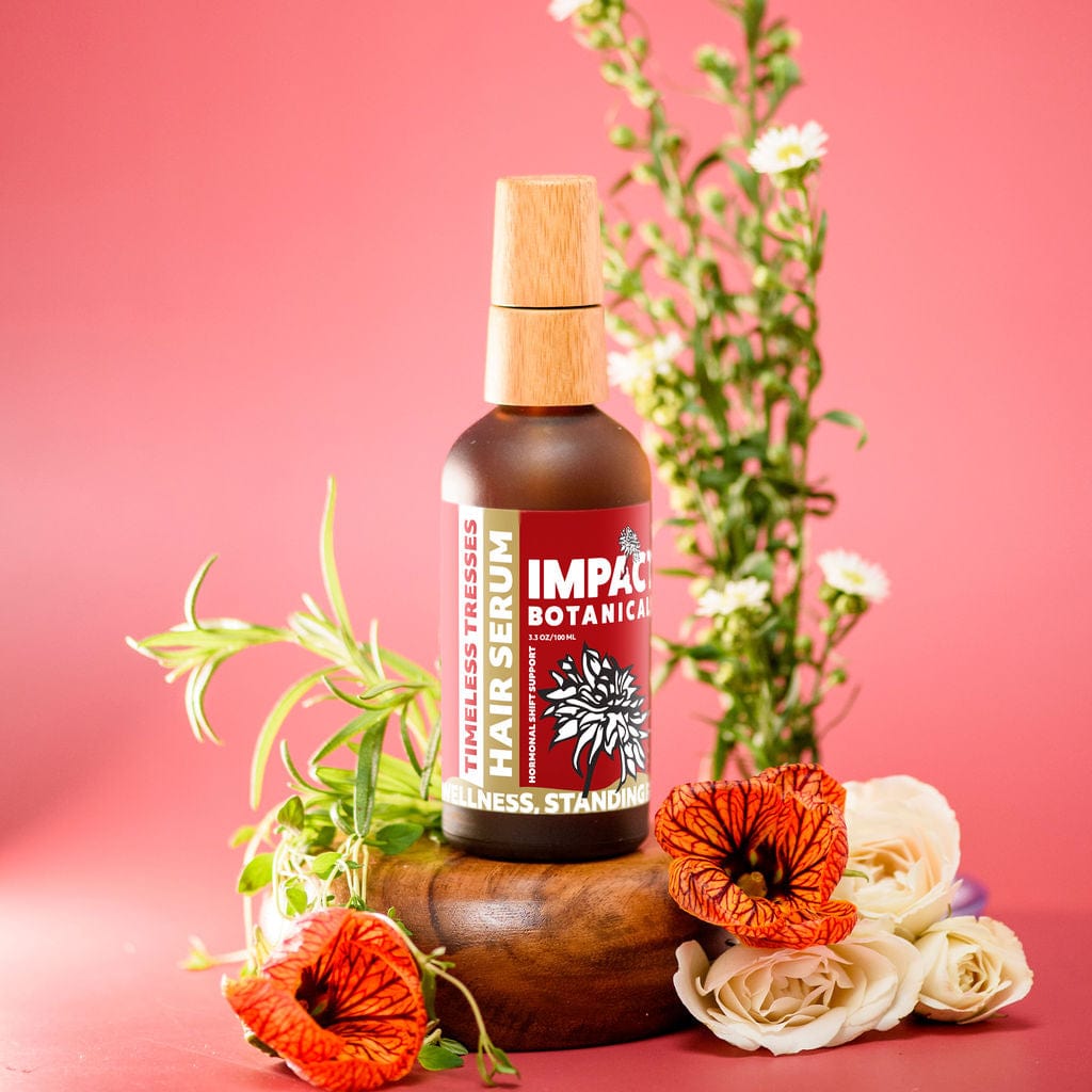 A bottle of Impact Botanicals Timeless Tresses Hair Serum stands on a wooden platform surrounded by vibrant flowers and rosemary sprigs against a pink background.