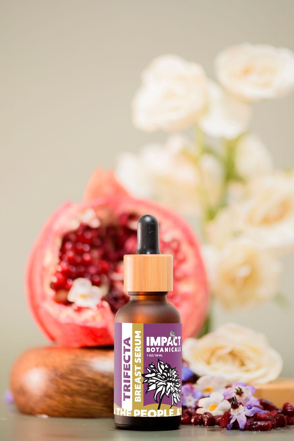 A bottle of Trifecta Breast Serum by Impact Botanicals stands on a wooden surface with a halved pomegranate behind it and white and purple flowers in the foreground.