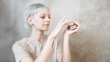 Woman holds a small jar of purple and gold skincare product in her hands, examining it closely.