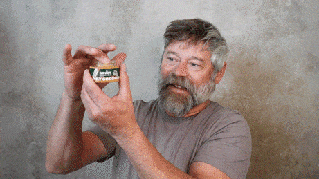 A bearded man examines a jar of "Impact Wellness Honey Goodness" in front of a textured gray wall.