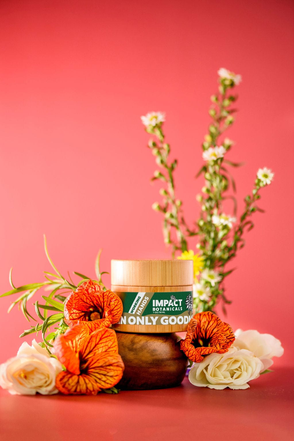 A bamboo jar of Impact Botanicals "Working Hands" cream sits on a wooden pedestal, surrounded by vibrant orange and white flowers against a bright pink backdrop.